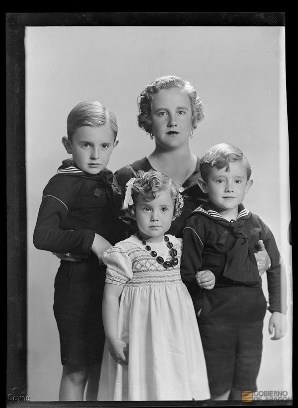 Retrato en plano americano de mujer de mediana edad seria detrás de dos niños y una niña con vestido y collar